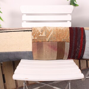 May include: A rectangular decorative pillow with a patchwork design. The pillow features sections of cream, gray, and patterned fabrics in shades of beige, brown, and red. It is placed on a white chair.