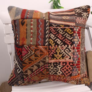 May include: A square, patchwork pillow with a variety of geometric patterns and colors. The pillow features a mix of warm tones, including red, orange, brown, and beige. The pillow is placed on a white chair.