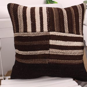 May include: A square decorative pillow with a patchwork design. The pillow features alternating vertical stripes in shades of brown and cream. The bottom of the pillow is a solid dark brown color. The pillow is made of a woven fabric.
