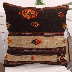 May include: A decorative square pillow with a woven design. The pillow features a dark brown background with geometric patterns in orange, red, and yellow. The lower section has horizontal stripes in beige and brown. Placed on a white chair.