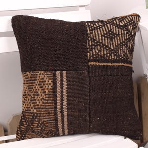 May include: Square cushion with a patchwork design. The cushion features a combination of dark brown and tan woven fabrics. The textures include solid, striped, and patterned sections, creating a rustic aesthetic. The cushion is placed on a white chair.