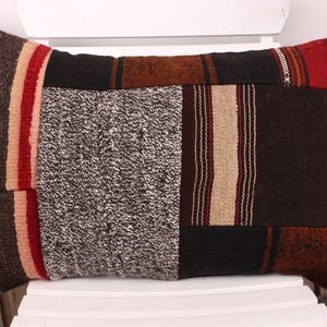 Puede incluir: Cojín decorativo rectangular con diseño de patchwork. El cojín presenta una mezcla de texturas y colores, incluyendo marrón, rojo, beige y gris. El diseño incluye rayas y secciones texturizadas, creando una estética rústica.