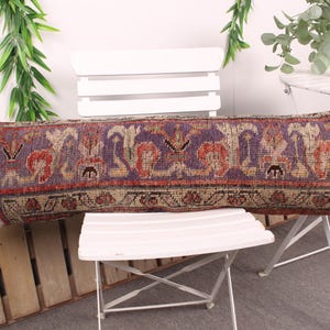 May include: A rectangular decorative pillow with a patterned design. The pillow features a purple background with red, orange, and beige floral motifs. The pillow is placed on a white chair.