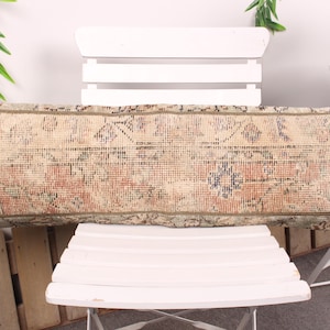Puede incluir: Cojín rectangular beige y rojo estampado. El cojín tiene un diseño de alfombra vintage con un aspecto desgastado. Se coloca sobre una silla blanca. Es un artículo decorativo.
