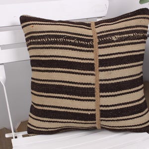 May include: Square cushion with a beige and dark brown striped design. It features a vertical beige stripe down the middle. The cushion is placed on a white chair.