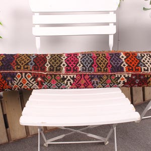 May include: A long, rectangular pillow with a vibrant, patterned design. The pillow features a mix of colors including red, pink, blue, and orange, with geometric and floral motifs. It rests on a white chair.