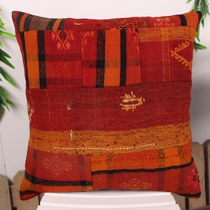 May include: Square decorative pillow with a patchwork design in shades of red, orange, and black. The pillow features a textured, woven fabric with geometric patterns and a rustic aesthetic. The pillow is placed on a white surface.
