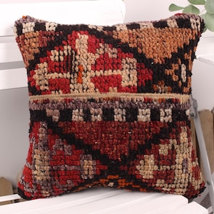 May include: Square decorative pillow with a geometric pattern in shades of red, black, tan, and brown. The pillow has a textured, woven appearance and is placed on a white chair.
