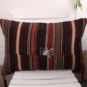May include: Rectangular pillow with a striped pattern in shades of brown, red, and beige. The pillow is placed on a white chair. The pillow has a distressed area with visible threads.