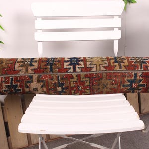 May include: A long, rectangular pillow with a patterned design in shades of red, blue, brown, and beige. The pillow is placed on a white chair, showcasing its intricate, geometric pattern. The pillow's design suggests a vintage or ethnic style.