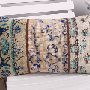 Peut inclure: Coussin rectangulaire décoratif avec un motif beige, bleu et marron. Le coussin présente un panneau central avec des motifs floraux bleus, flanqué de bordures à motifs turquoise et marron. Le coussin est posé sur une chaise blanche.
