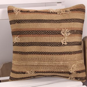 Könnte beinhalten: Quadratisches Kissen mit einem gewebten Streifendesign in Beige-, Braun- und Cremetönen. Das Kissen hat einen Used-Look mit Bereichen mit offenem Gewebe. Das Kissen liegt auf einer weißen Oberfläche.