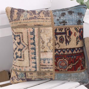 May include: Square patchwork pillow with a vintage rug design. The pillow features a mix of beige, blue, and red colors with geometric and floral patterns. The pillow is placed on a white chair.