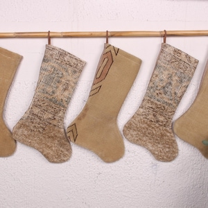 Op de afbeelding: Vijf beige kerstsokken hangen aan een houten roede. Twee sokken hebben patronen in bruin- en blauwtinten. De andere drie sokken zijn effen beige. De sokken zijn gemaakt van een textuurstof.