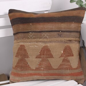 May include: Square decorative pillow with a woven design in shades of brown, tan, and orange. The pillow features a geometric pattern of triangles and horizontal stripes. The pillow is placed on a white chair.