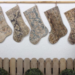 May include: Five decorative Christmas stockings made from repurposed rugs. The stockings are hanging from a wooden dowel rod against a white wall. The stockings are beige with brown and blue patterns.