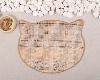 May include: A beige cat-shaped pet mat with a textured surface. The mat is designed to look like a cat's face with a brown outline around the ears and eyes.