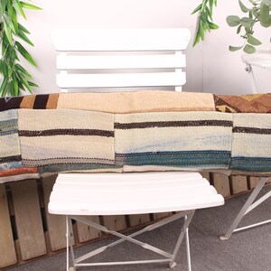 Op de afbeelding: Een lang, rechthoekig kussen met een patchwork-ontwerp. Het kussen heeft delen van beige, bruine en blauwe stof met gestreepte en patroon details. Het rust op een witte stoel en tafel, wat duidt op een decoratief woonaccessoire.