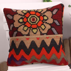 May include: A decorative pillow with a geometric design. The top features a floral pattern in shades of orange, tan, and black, while the bottom has a zig-zag pattern in red, orange, and brown. The pillow is placed on a white chair.