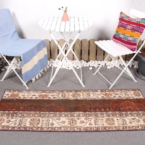 Op de afbeelding: Een bruin en crèmekleurig tapijt met een wit, bruin en blauw gestreept plaid op een witte vouwstoel. Een witte vouwtafel met een roze bloem in een witte vaas staat voor de stoel.