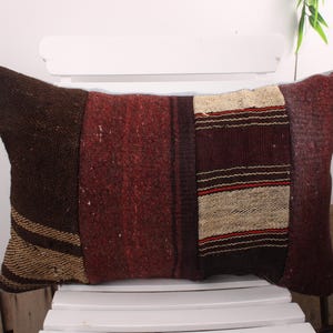 May include: Rectangular cushion with a patchwork design. The cushion features sections of brown, red, and beige fabric. The beige section has horizontal stripes with red accents. The cushion is placed on a white chair.