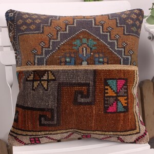 May include: Square decorative pillow with a patchwork design. The pillow features a mix of brown, orange, gray, and blue geometric patterns. The pillow is placed on a white chair, showcasing its intricate details and textures.
