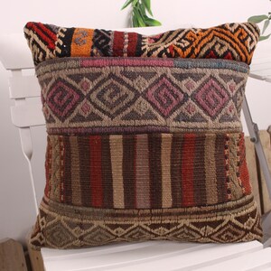 Puede incluir: Un cojín decorativo con un diseño de patchwork. El cojín presenta patrones geométricos en tonos marrones, rojos, naranjas y morados. El cojín es rectangular y parece estar hecho de tela tejida. Se coloca sobre una silla blanca.