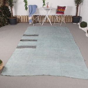 Puede incluir: Una alfombra grande, rectangular, de color azul claro con tres rayas marrones oscuras en un extremo. La alfombra está hecha de un material tejido y tiene un aspecto ligeramente desgastado. La alfombra se muestra sobre una superficie gris.