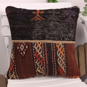 May include: A decorative pillow with a woven design. The top portion is dark brown with orange and green geometric shapes. The bottom section is brown with orange, red, and white patterns. The pillow is square and appears to be handmade.