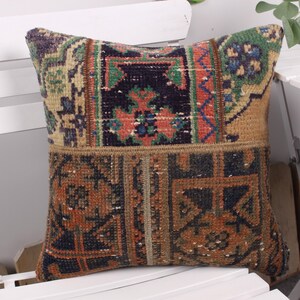 May include: Square decorative pillow with a patchwork design. The pillow features a mix of colors including brown, orange, green, and blue. The pillow is placed on a white chair.