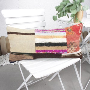 May include: A long, rectangular patchwork cushion with a beige background and colourful stripes. The cushion is made of different fabrics and has a brown, black and white striped pattern on one side. The cushion is sitting on a white chair.