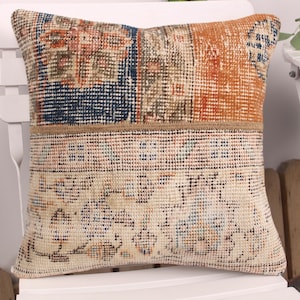 May include: A square pillow with a patchwork design. The top section features a mix of orange, navy, and brown patterns, while the bottom section has a cream-colored, patterned design. The pillow is placed on a white chair.