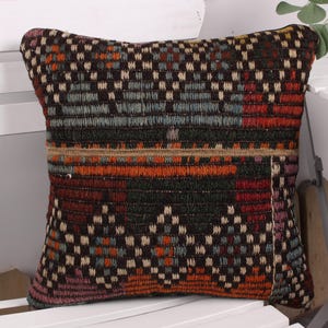 May include: A square decorative pillow with a woven, geometric pattern. The pillow features a mix of colors, including black, orange, red, and blue. The pillow is placed on a white chair.
