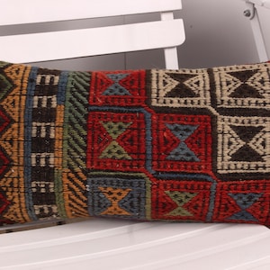Puede incluir: Cojín rectangular decorativo con un patrón geométrico tejido. El cojín presenta una mezcla de colores que incluyen rojo, azul, dorado y negro. El cojín está sobre una silla blanca.