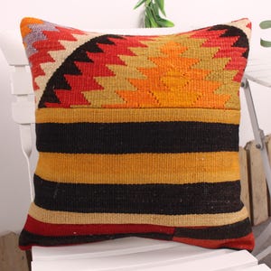 May include: A decorative pillow with a geometric design. The top section features a zigzag pattern in red, orange, yellow, and beige. The lower section has horizontal stripes in black, yellow, and red. The pillow is placed on a white chair.