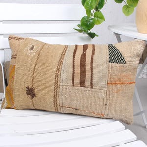 May include: Rectangular beige pillow with a patchwork design. The pillow features various textures and patterns, including brown stripes, geometric shapes, and embroidered details. It rests on a white chair.