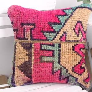 Puede incluir: Un cojín cuadrado decorativo con un patrón geométrico vibrante. El cojín presenta una mezcla de tonos rosa, beige, turquesa y morado. El cojín está hecho de un material texturizado y está colocado sobre una silla blanca.