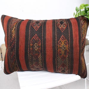 May include: Rectangular pillow with a woven design. The pillow features vertical stripes in shades of red and black, with intricate geometric patterns in the center of the stripes. The pillow is placed on a white chair.