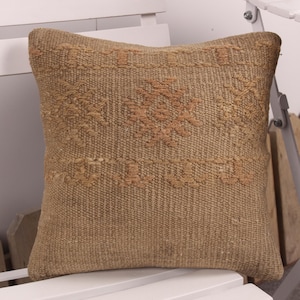 May include: A square, woven pillow in shades of tan and beige. The pillow features a textured surface with a detailed, geometric pattern in a lighter tan color. The pillow is placed on a white surface.