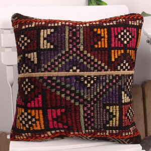 May include: Square decorative pillow with a woven, geometric pattern in shades of red, orange, purple, and black. The pillow is placed on a white chair, showcasing its intricate design and vibrant colors. The pillow's dimensions are not visible.