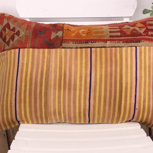 Puede incluir: Cojín rectangular con un estampado de rayas en tonos amarillos, marrones y beige. La sección superior presenta un diseño geométrico en rojo, naranja y marrón. Una raya vertical azul acentúa el frente.