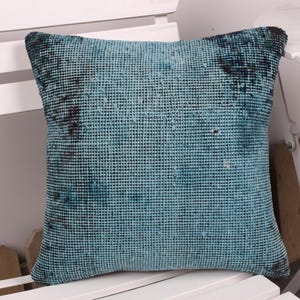 May include: Square turquoise pillow with a textured, woven design. The pillow has a faded appearance with darker blue accents. The pillow is placed on a white chair.