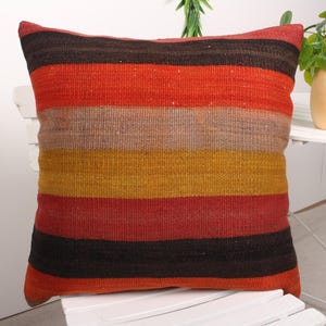 May include: A square decorative pillow with horizontal stripes in shades of red, brown, and gold. The pillow is made of a woven fabric and rests on a white chair. The pillow's design adds a touch of warmth and texture to any living space.