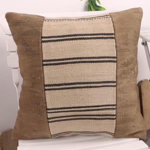 May include: Square decorative pillow with a textured, brown fabric. The center panel is a lighter beige with horizontal black stripes. The pillow is placed on a white chair, suggesting a home decor theme.