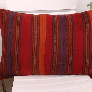 May include: Rectangular pillow with a striped pattern in shades of red, orange, and purple. The pillow is made of woven fabric and is resting on a white chair. The pillow's dimensions are not visible.