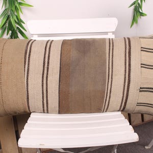 Puede incluir: Cojín rectangular beige con rayas verticales marrones y un panel central marrón. El cojín está hecho de tela tejida y descansa sobre una silla blanca. El diseño es sencillo y rústico, adecuado para la decoración del hogar.