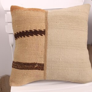 May include: Square decorative pillow with a two-tone design. One side is a light beige color, and the other side is a tan color with a dark brown geometric pattern. The pillow is made of a woven fabric.