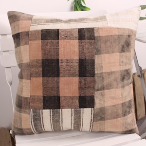 May include: Square decorative pillow with a patchwork design. The pillow features a plaid pattern in shades of brown, tan, and cream. The pillow is placed on a white chair, suggesting a home decor theme.