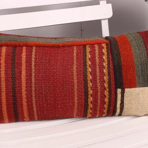 May include: Rectangular decorative pillow with a patchwork design. The pillow features a red background with vertical stripes in shades of orange, brown, and green. The ends have a beige and brown pattern. The pillow is placed on a white chair.