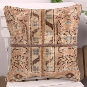 May include: Square decorative pillow with a vintage rug design. The pillow features a beige, brown, and blue color palette with a geometric pattern. The pillow is placed on a white chair.